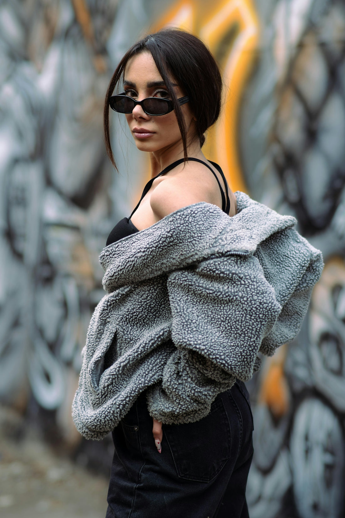 Woman in fuzzy jacket and sunglasses poses against graffiti wall